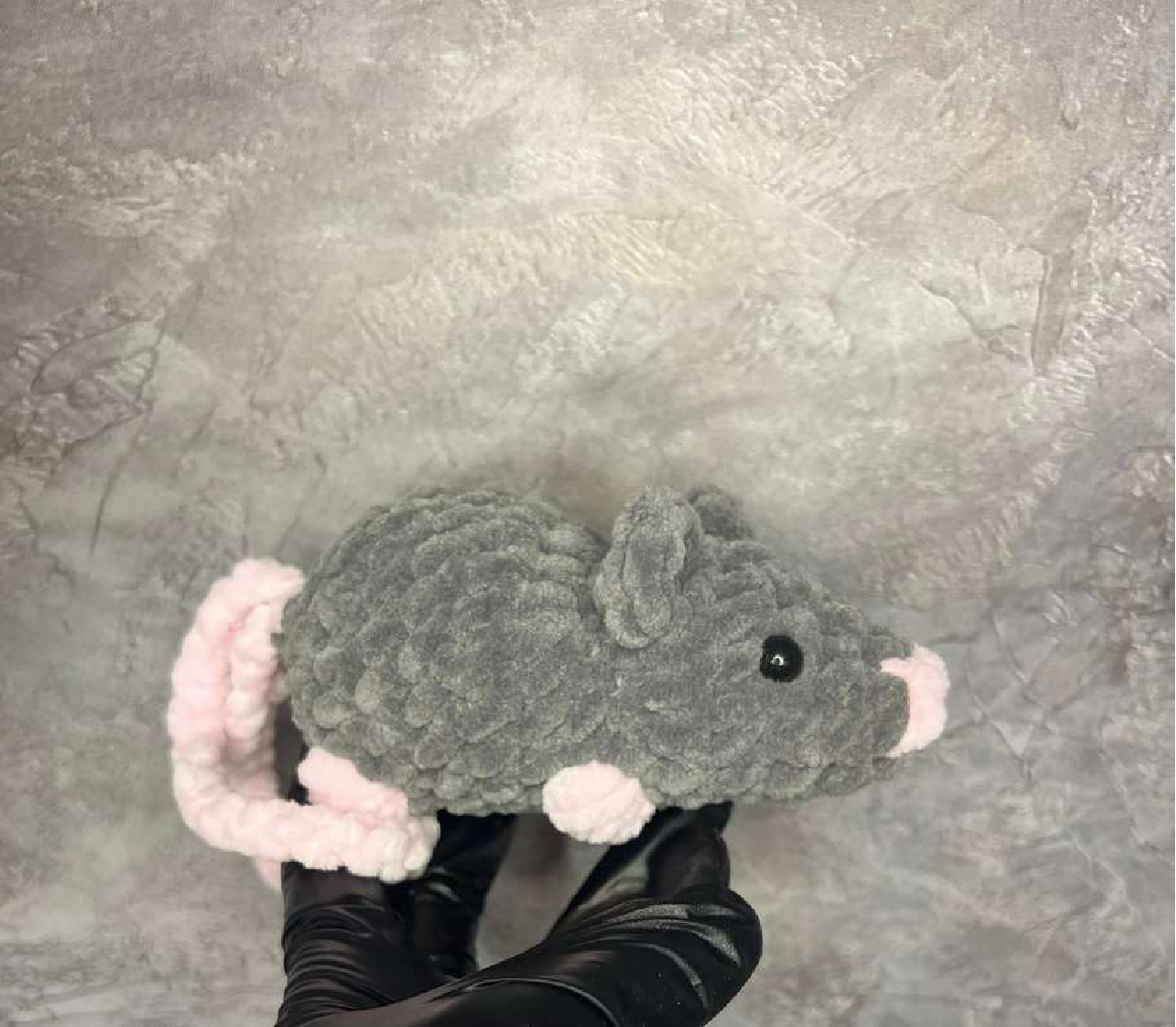 A standard crocheted grey mouse with pink ears, feet, and nose, lying on a grey textured surface.