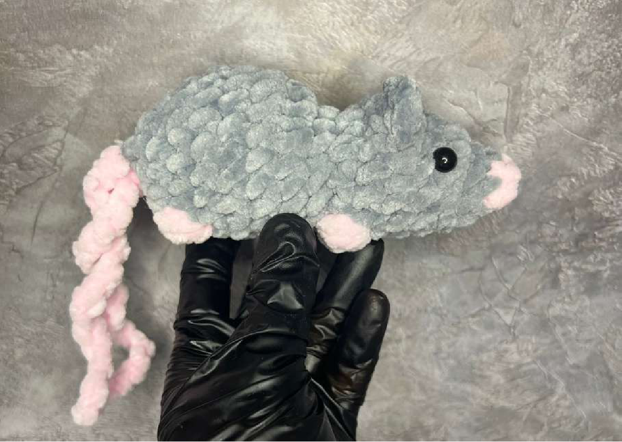 A side view of the light grey crocheted mouse, showing its body, pink tail, and smaller ears.