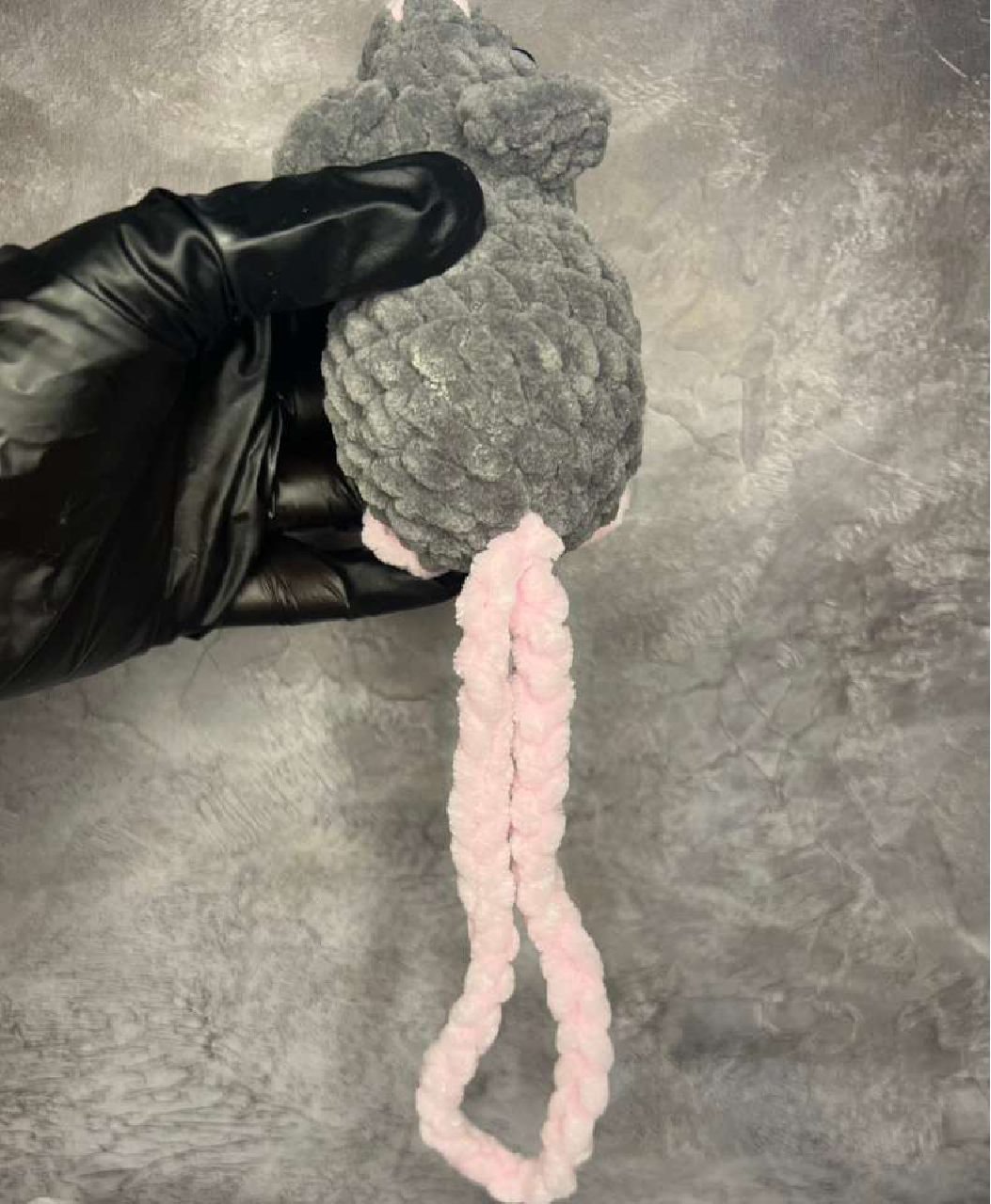 A crocheted grey mouse from behind, showing its pink tail.