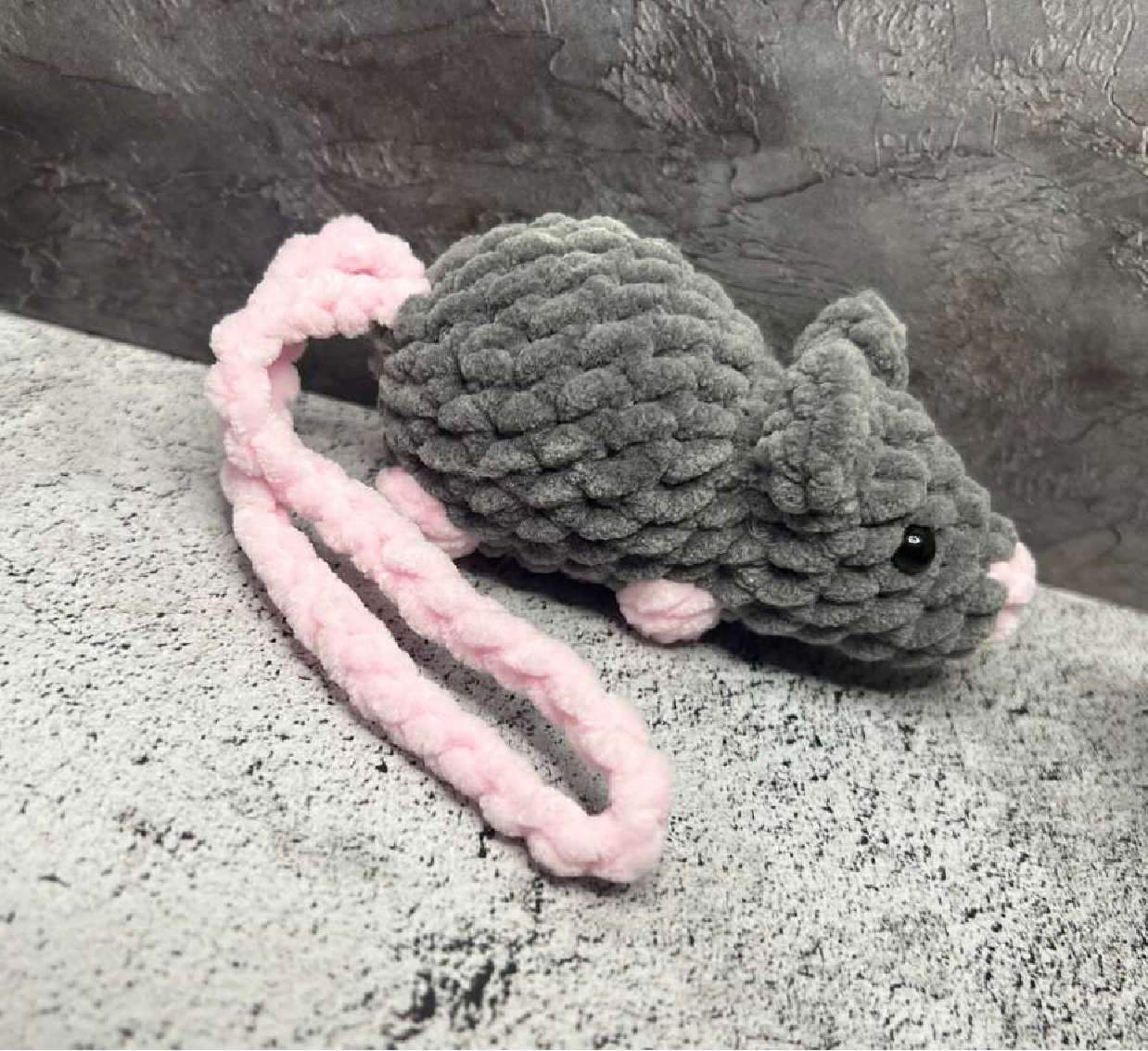 A completed crocheted grey mouse with a long pink tail, lying on a grey textured surface.