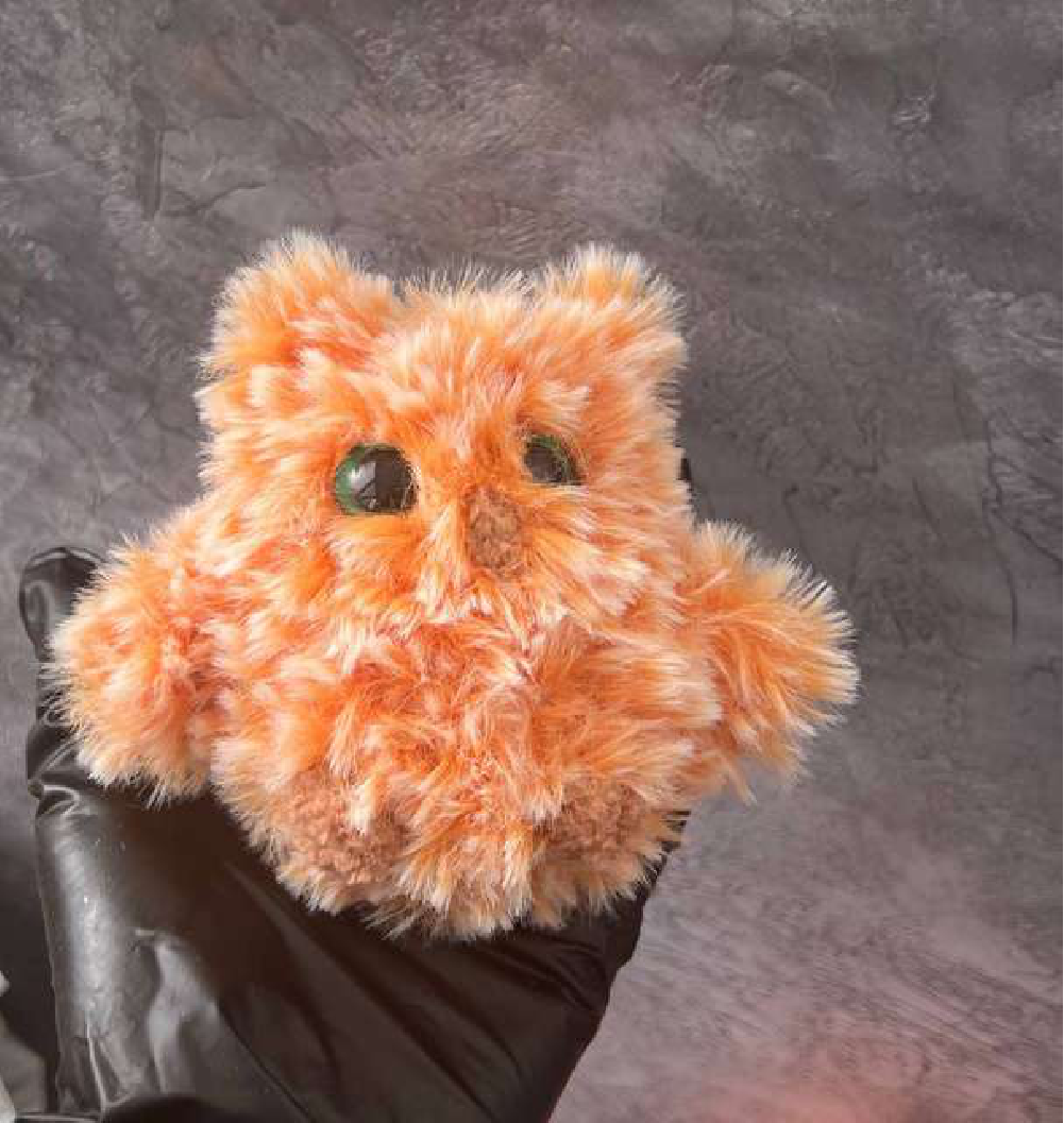 A fluffy, orange, small owl toy held in a hand wearing a black glove.